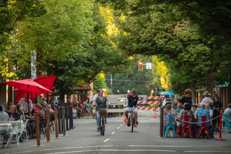 Three bikers with one waving ride by groups of people dining in parklets in a blocked off pedestrian only street
