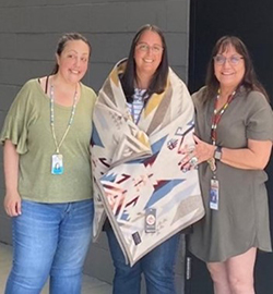 Three women stand together, the woman in the middle has been given a blanket