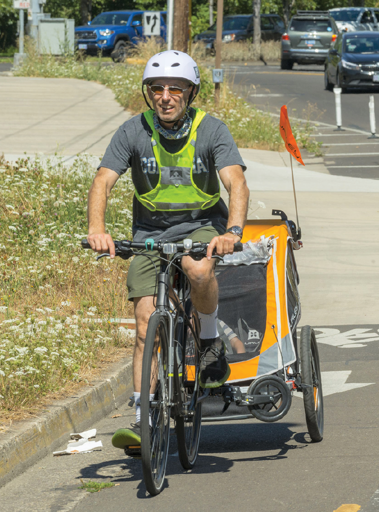 A man wearing a reflective vest riding a bicycle towing a passenger trailer with a child inside.