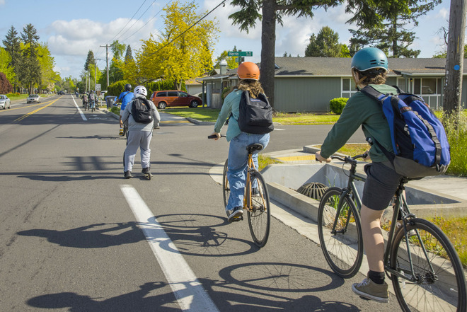 Several people shown riding their bikes in a bike lane next to a multi-lane road in a residential area