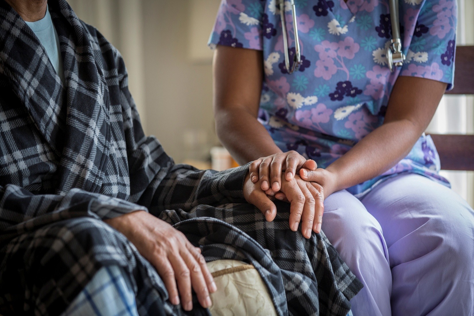 A nurse wearing floral scrubs holds the hand of an older patient.
