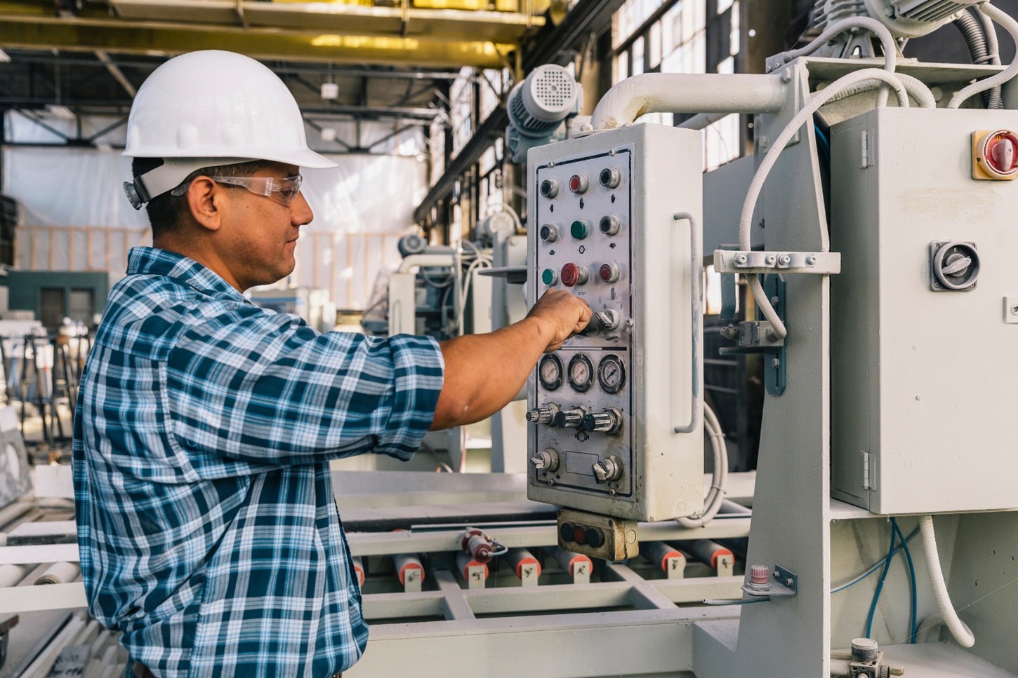 A worker wearing a white hardhat and safety glasses turns a knob on a panel.