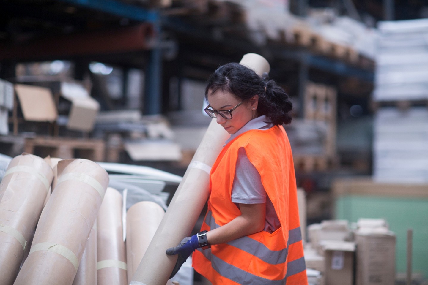 A worker wearing an orange vest lifts a large roll of brown paper.