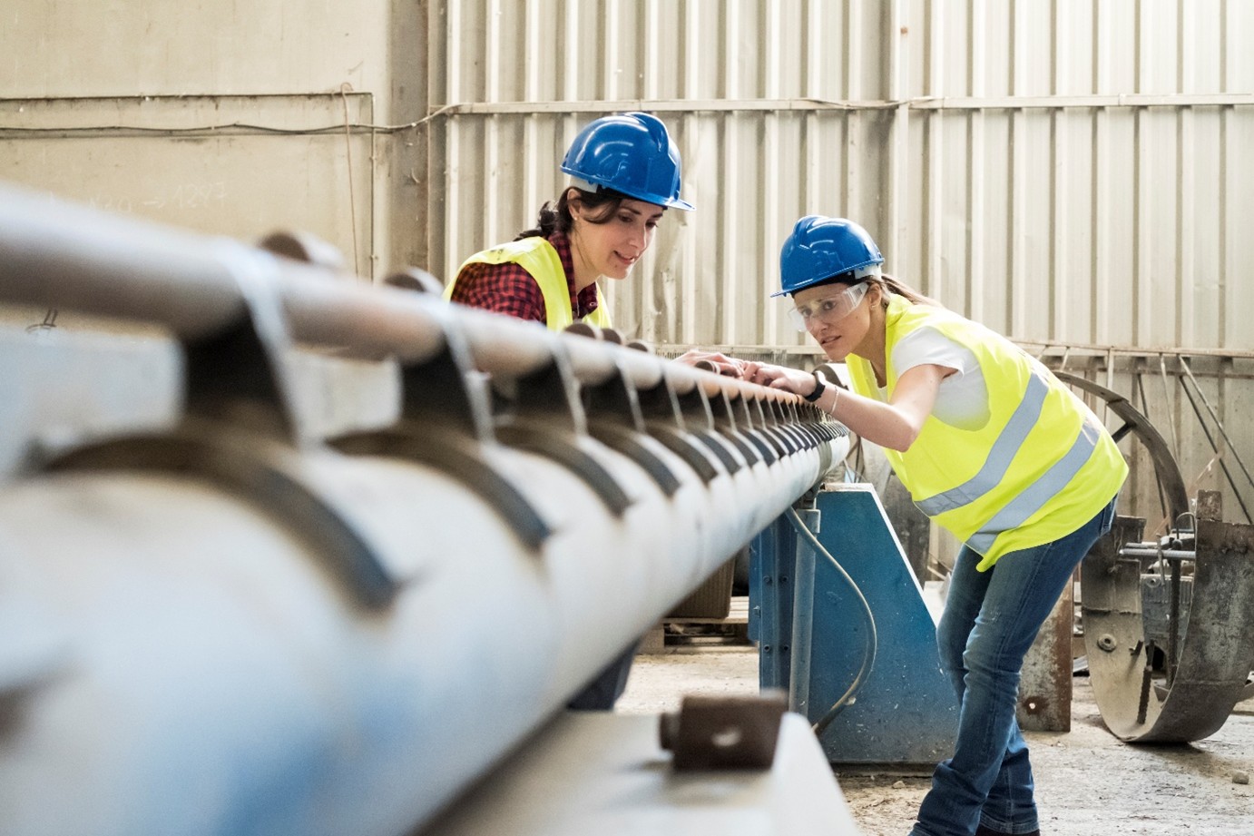 2 workers looking at machinery together. They are both wearing hardhats, and one has on protective googles.