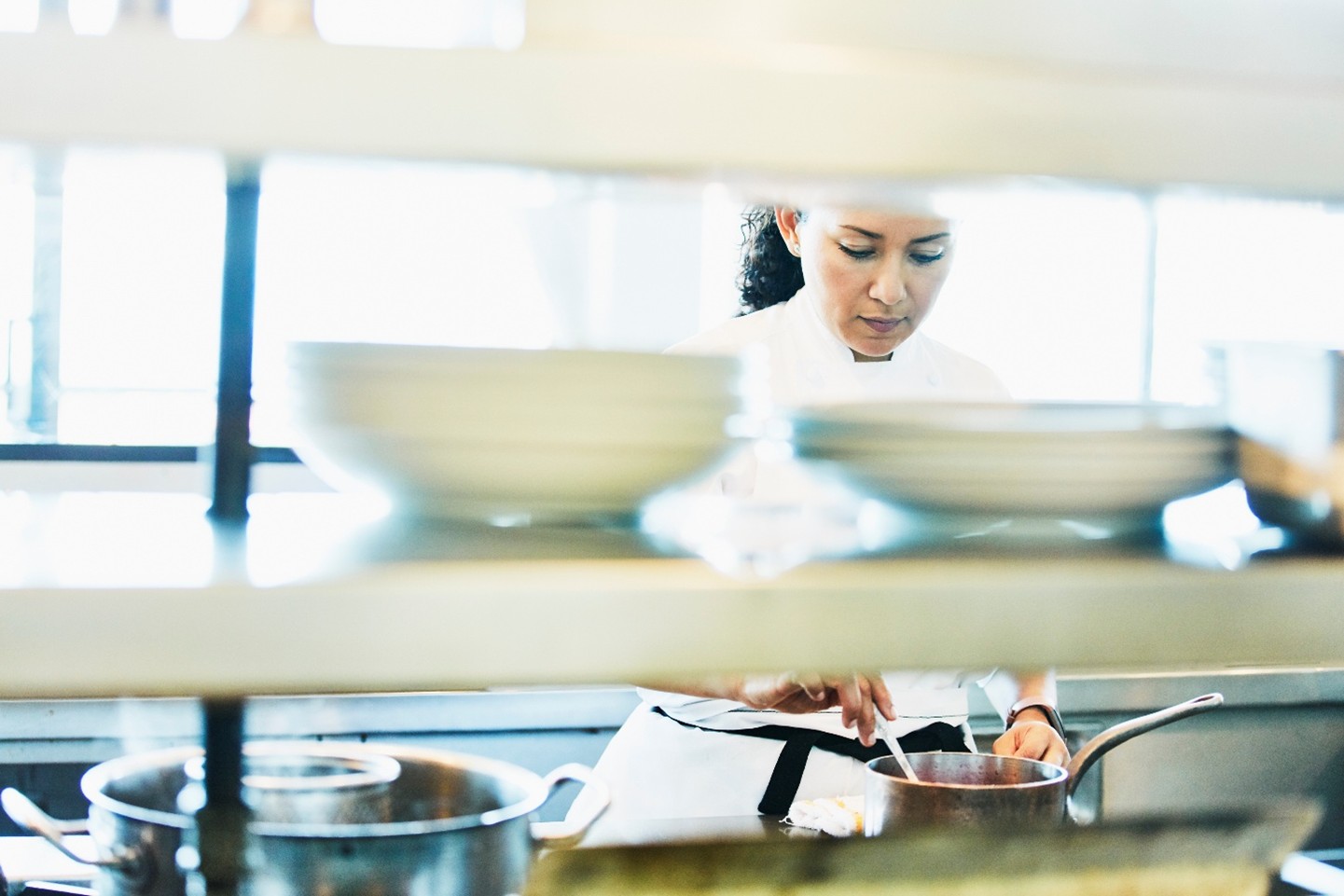 A chef inside a kitchen stirring food in a metal pot.