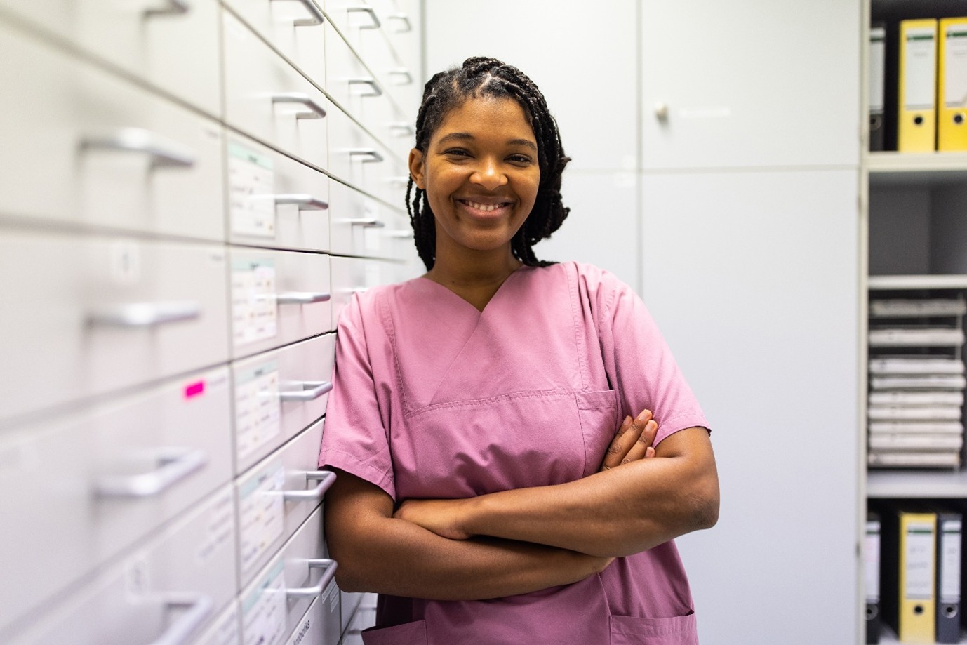 A smiling person wearing scrubs stands with arms crossed next to filing cabinets.