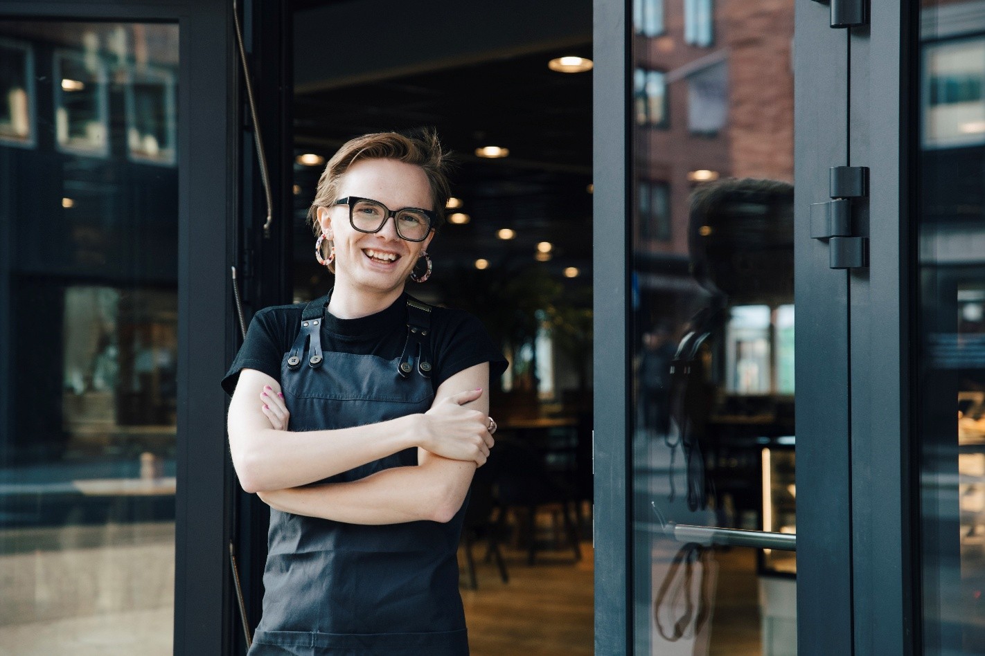 A young worker wearing a black apron stands in front of an open cafe door