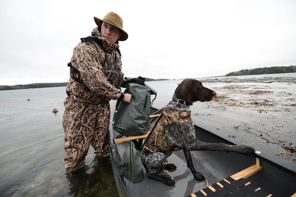 Old Town Canoe image of a waterfowl hunter in a canoe prepping to get underway with a dog. Both are wearing life jackets.